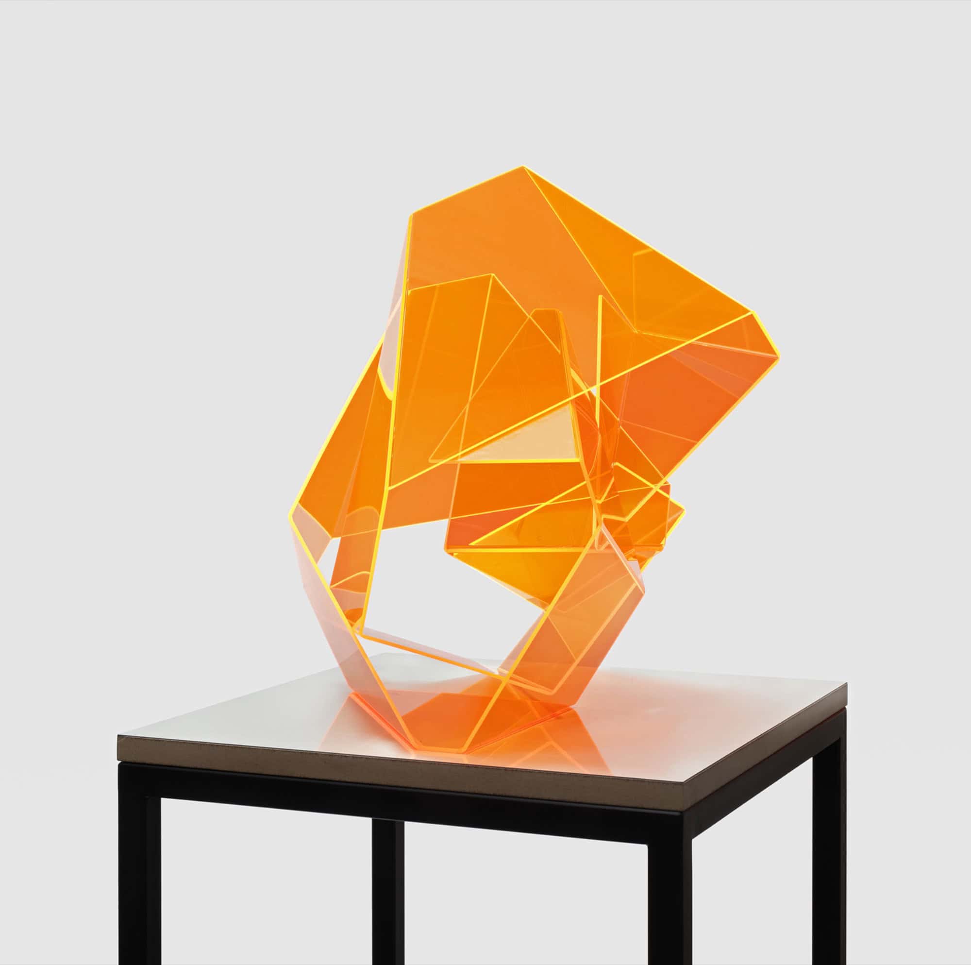 Leuchtend orangefarbene Skulptur aus gefaltetem fluoreszierendem Acrylglas auf einem weißen Sockel mit schwarzem Metallgestell vor grauer Wand.