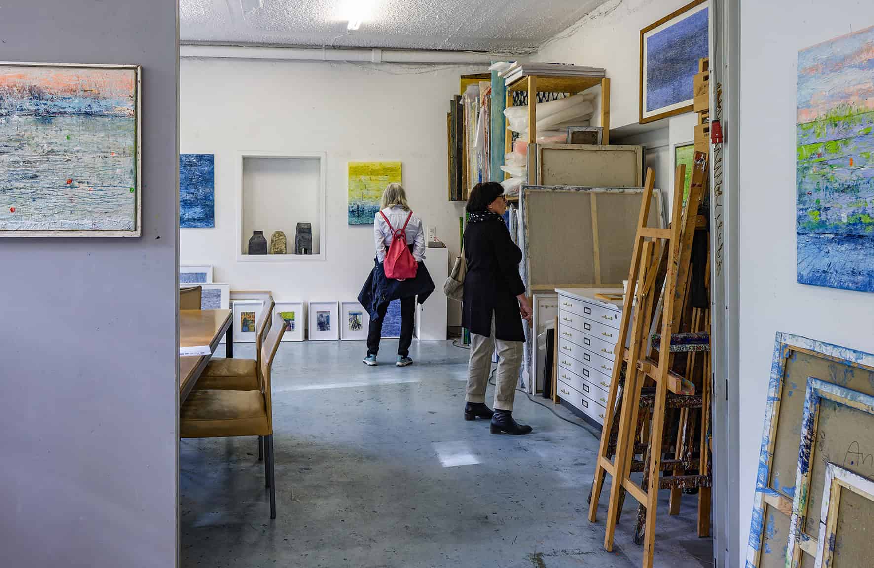 Zwei Besucherinnen betrachten Bilder in einem Atelier voller gerahmter Werke und Staffeleien