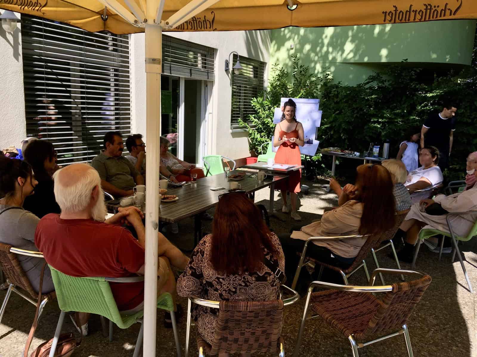 Nachbarinnen und Nachbarn sitzen unter Sonnenschirmen an Tischen zusammen, eine Frau spricht zur Runde.
