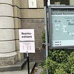 Rosafarbenes Schild mit der Aufschrift Bezirkswahlamt und den Öffnungszeiten neben einem Übersichtsplan des Bezirksamtes Reinickendorf an der Teichstraße.