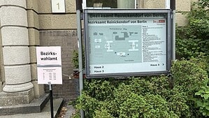Rosafarbenes Schild mit der Aufschrift Bezirkswahlamt und den Öffnungszeiten neben einem Übersichtsplan des Bezirksamtes Reinickendorf an der Teichstraße.