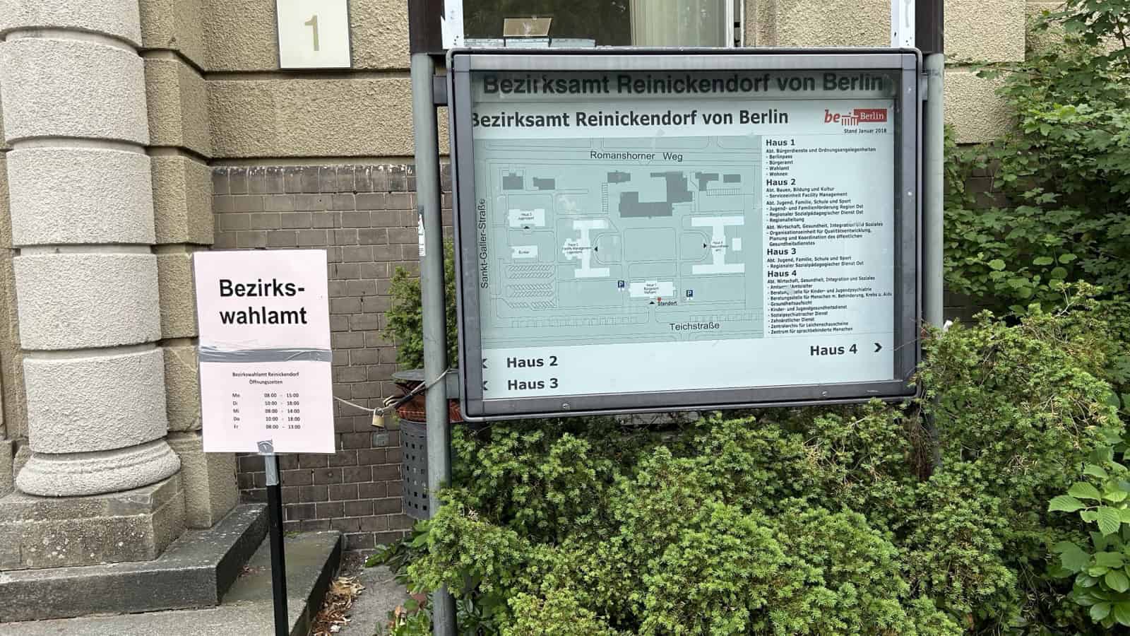 Rosafarbenes Schild mit der Aufschrift Bezirkswahlamt und den Öffnungszeiten neben einem Übersichtsplan des Bezirksamtes Reinickendorf an der Teichstraße.