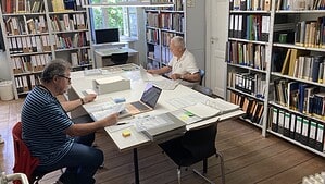 Zwei Männer sitzen an einem großen weißen Tisch in einem Bibliotheksraum und arbeiten mit Dokumenten, Mappen und einem Laptop. Ringsum stehen Regale mit Aktenordnern und Büchern, durch das Fenster fällt Tageslicht.