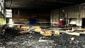 Blick in eine ausgebrannte Sporthalle: Der Boden ist von schwarzen Trümmern und Asche bedeckt, die Decke ist verrußt und teilweise heruntergefallen, an der Wand steht noch eine blaue Matte.