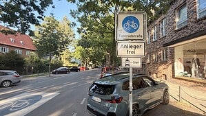 Ein Verkehrszeichen "Fahrradstraße" steht an einer Straße. Am Straßenrand parken Autos. Die Straße ist von grünen Laubbäumen gesäumt.