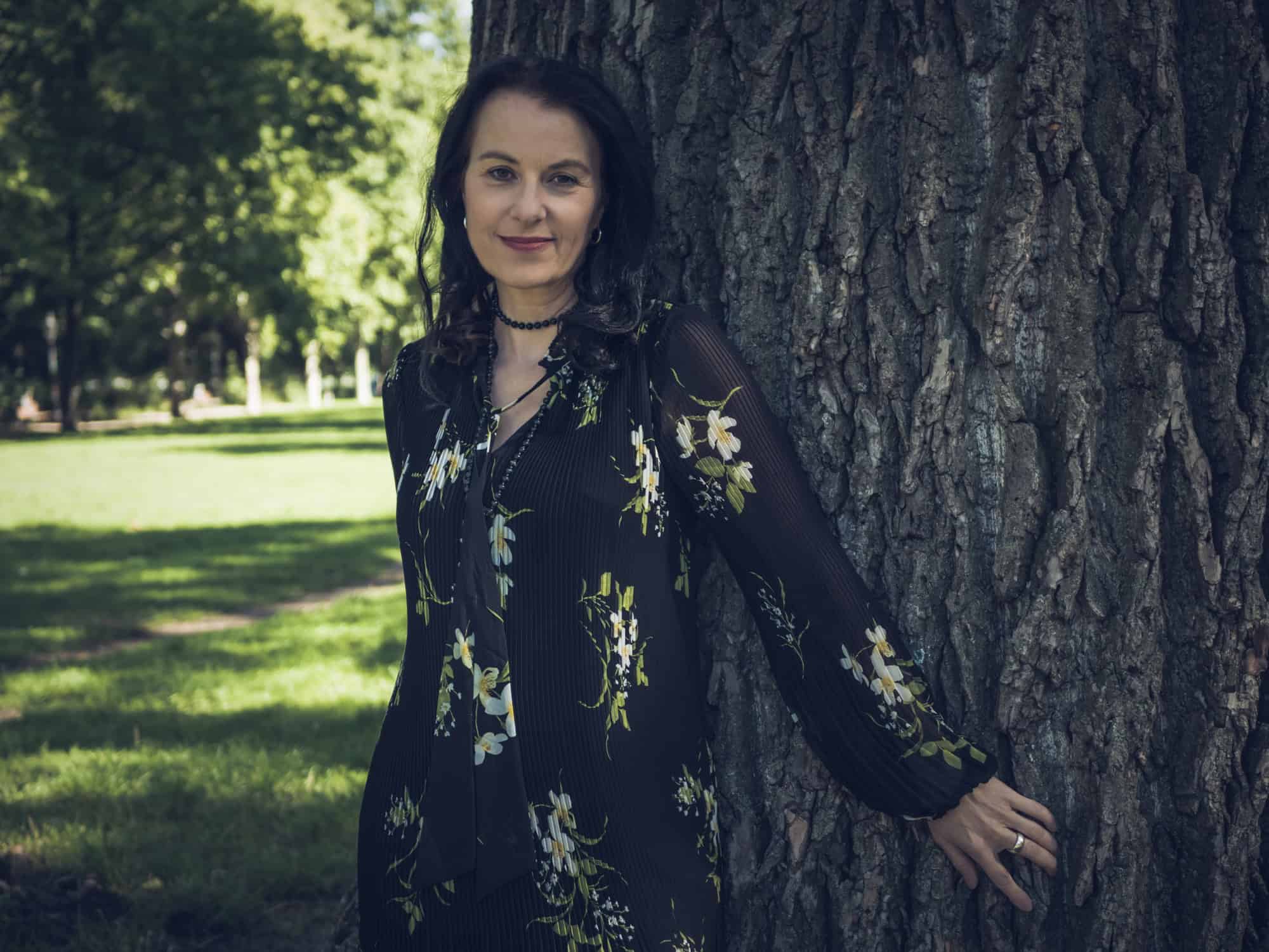 Autorin Elena Eden steht in einem sonnigen Park an einen dicken Baumstamm gelehnt und blickt in die Kamera.
