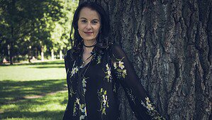 Autorin Elena Eden steht in einem sonnigen Park an einen dicken Baumstamm gelehnt und blickt in die Kamera.