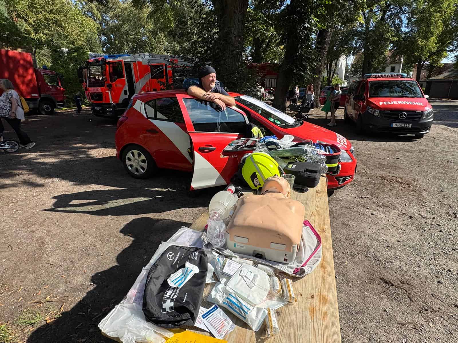 Auf einer Holzbank liegen eine Übungspuppe zur Wiederbelebung, ein Beatmungsbeutel, ein Feuerwehrhelm und Erste-Hilfe-Material. Dahinter stehen ein rotes Einsatzfahrzeug mit geöffneter Tür, ein Löschfahrzeug und ein Feuerwehr-Transporter.