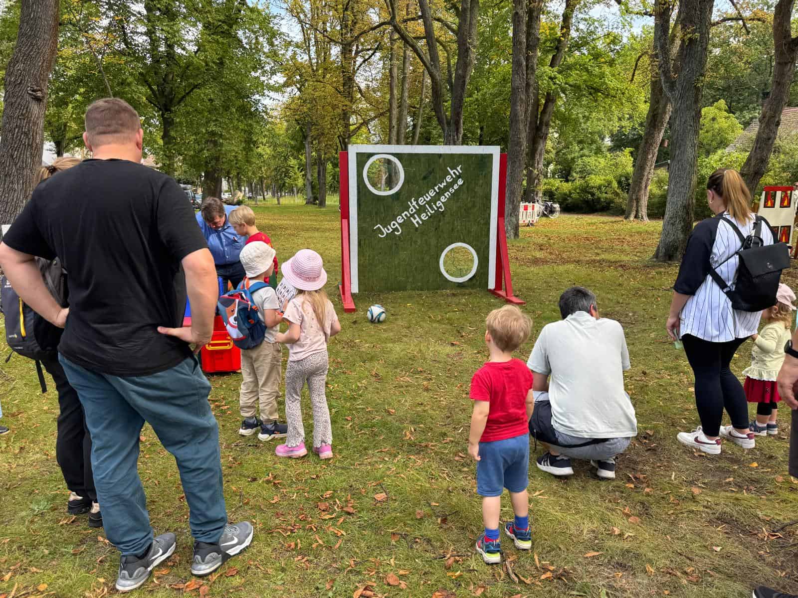 Kinder stehen mit einem Ball vor einer grünen Torwand mit der Aufschrift Jugendfeuerwehr Heiligensee, daneben schauen Erwachsene zu. Die Wand steht auf einer Wiese unter hohen Bäumen.