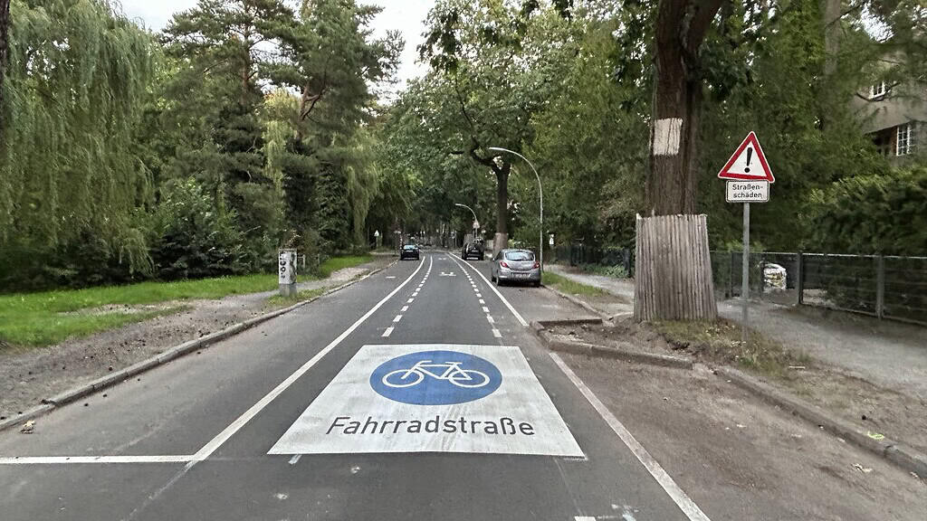 Blick über den Edelhofdamm mit dem Fahrbahnpiktogramm Fahrradstraße und einem Warnschild Straßenschäden.