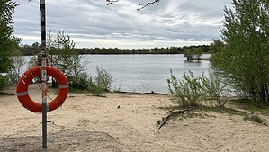 Rettungsring an einem Pfosten am Sandufer des Flughafensees in Berlin-Tegel, dahinter der See und bewaldete Ufer.