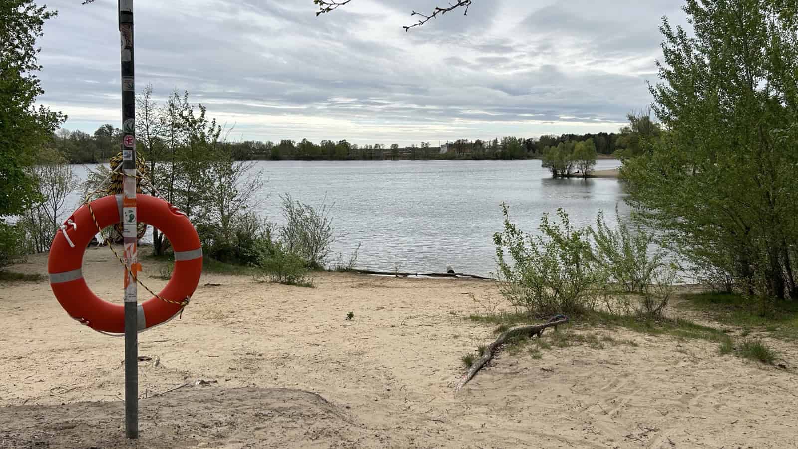 Rettungsring an einem Pfosten am Sandufer des Flughafensees in Berlin-Tegel, dahinter der See und bewaldete Ufer.