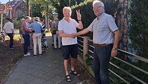 Zwei Männer geben sich auf einem Gehweg die Hand, einer zeigt das Victory-Zeichen, im Hintergrund eine Gruppe von Menschen.
