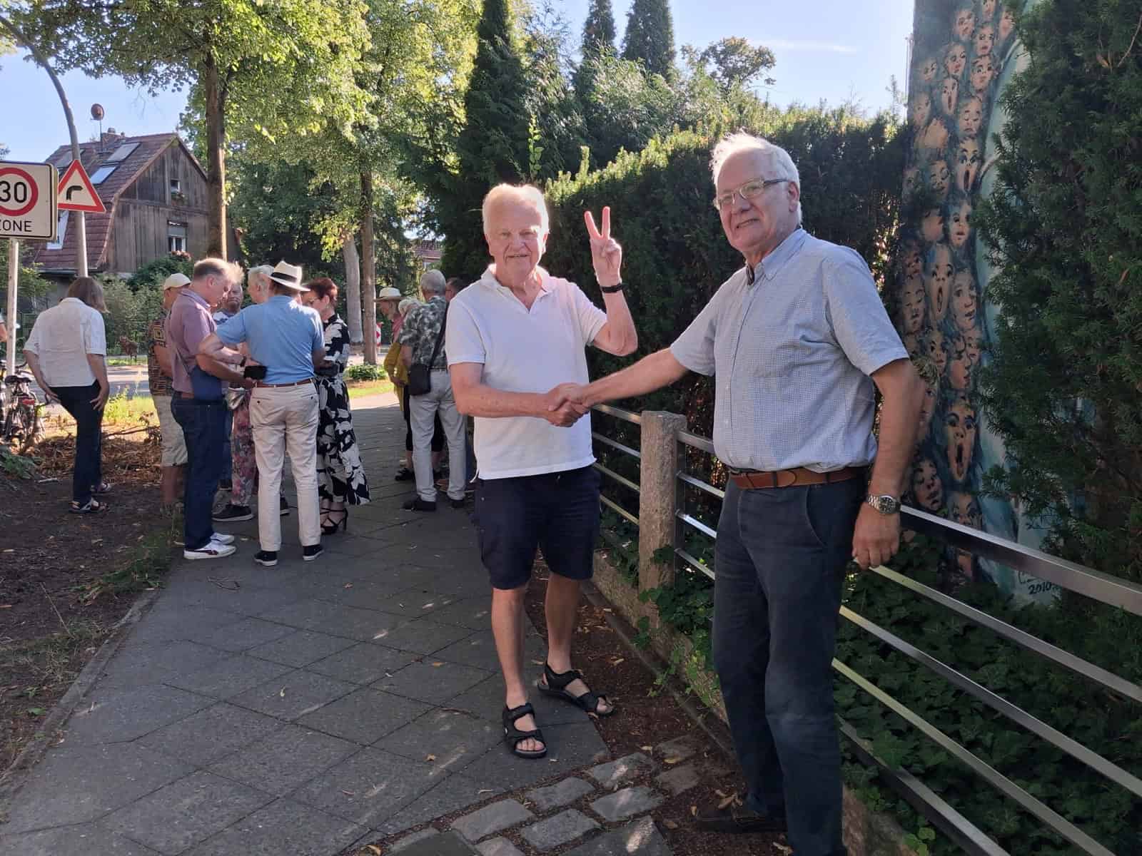 Zwei Männer geben sich auf einem Gehweg die Hand, einer zeigt das Victory-Zeichen, im Hintergrund eine Gruppe von Menschen.