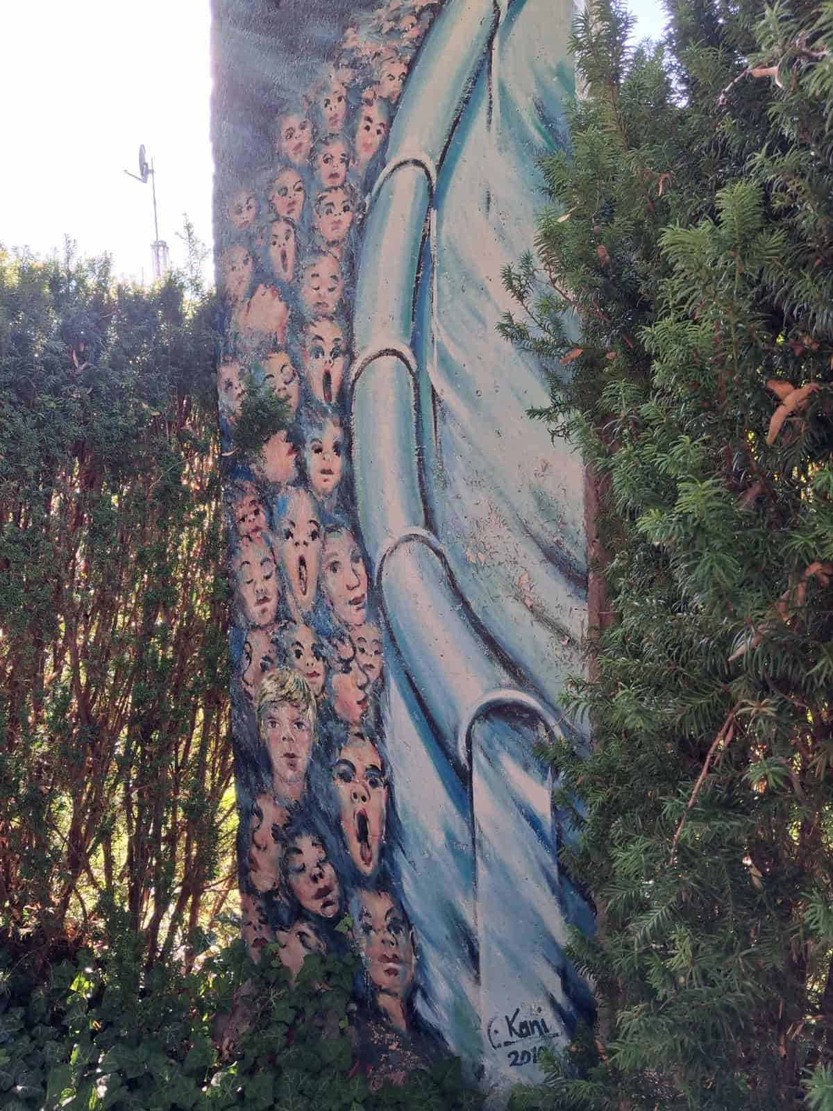 Ein erhaltenes Betonsegment der Berliner Mauer, bemalt mit zahlreichen Gesichtern, steht zwischen Hecken.
