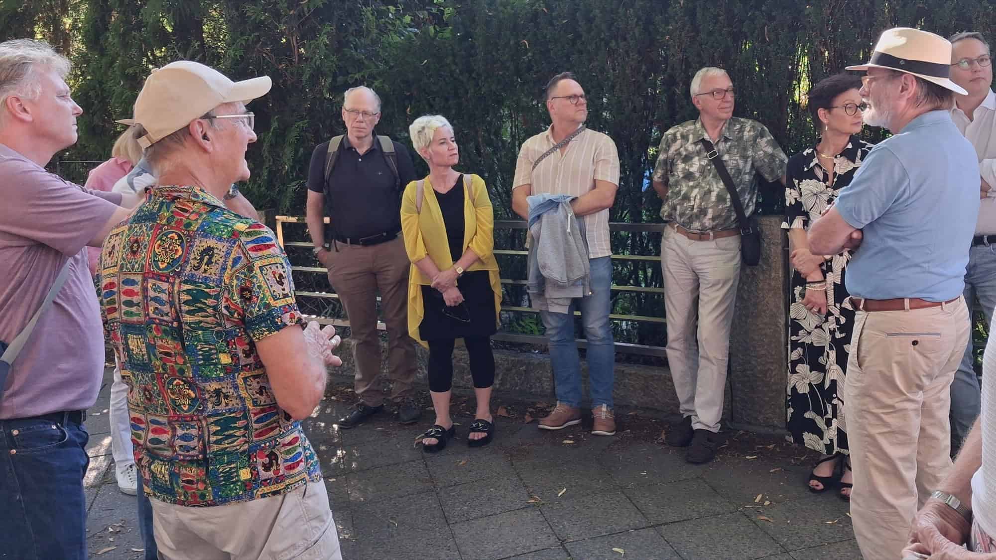 Eine Gruppe von etwa einem Dutzend Menschen steht im Halbkreis auf einem Gehweg vor einer hohen Hecke.