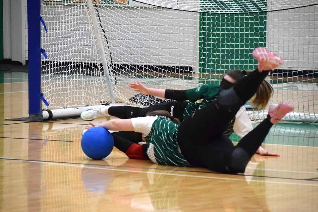 Zwei Spielerinnen in grünen Trikots und schwarzen Hosen liegen mit Augenbinden auf dem Hallenboden vor einem Tor, direkt vor ihnen ein blauer Ball.