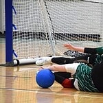 Zwei Spielerinnen in grünen Trikots und schwarzen Hosen liegen mit Augenbinden auf dem Hallenboden vor einem Tor, direkt vor ihnen ein blauer Ball.