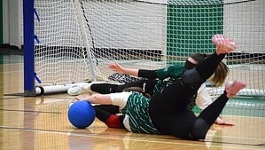 Zwei Spielerinnen in grünen Trikots und schwarzen Hosen liegen mit Augenbinden auf dem Hallenboden vor einem Tor, direkt vor ihnen ein blauer Ball.