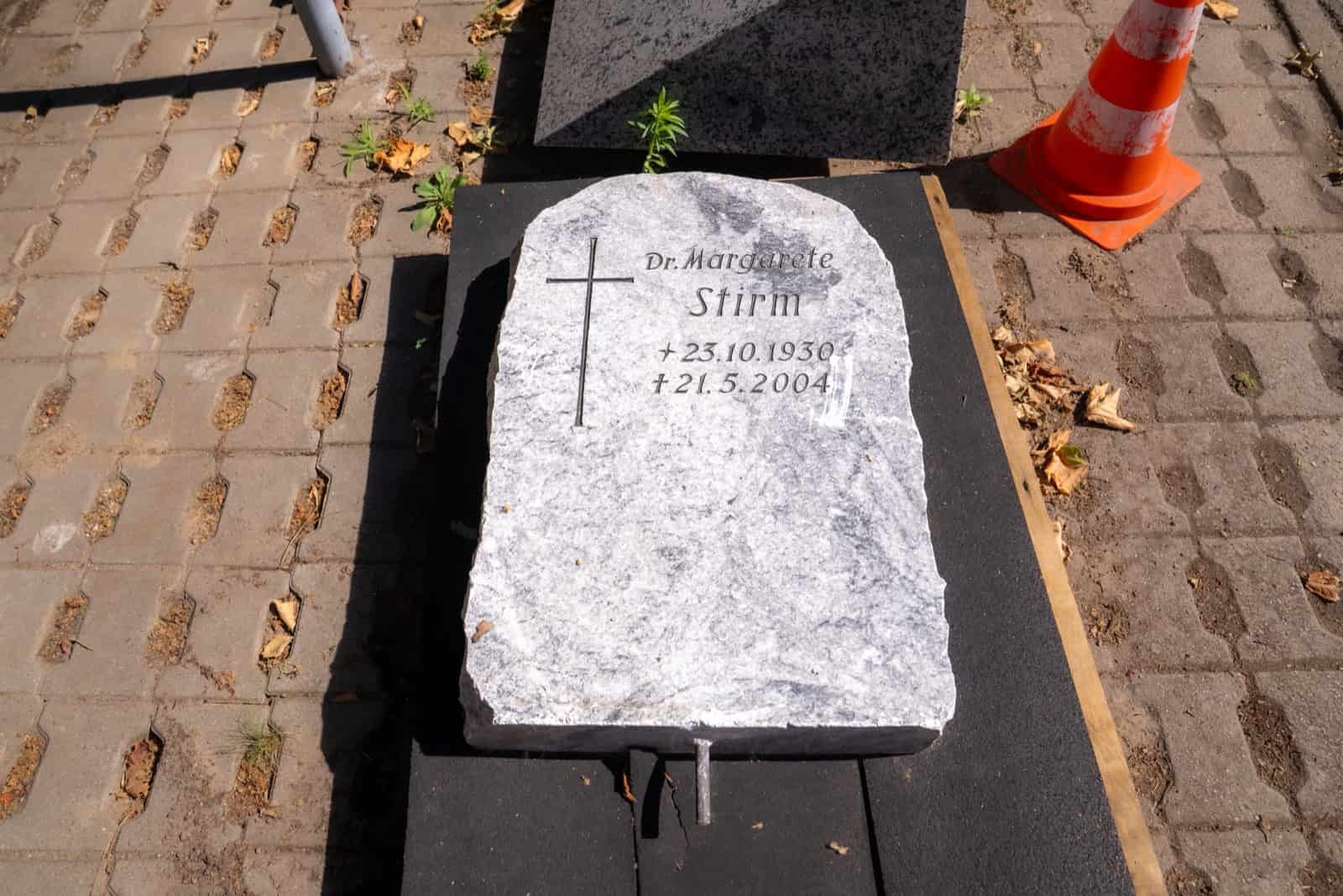 Ein heller, grob behauener Grabstein liegt flach auf einer Unterlage auf gepflastertem Boden. In den Stein sind ein Kreuz und die Inschrift „Dr. Margarete Stirm, + 23.10.1930, † 21.5.2004“ eingemeißelt, daneben steht ein orange-weißer Leitkegel.