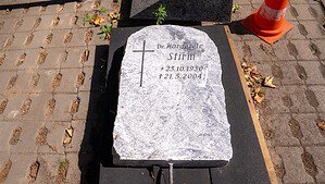 Ein heller, grob behauener Grabstein liegt flach auf einer Unterlage auf gepflastertem Boden. In den Stein sind ein Kreuz und die Inschrift „Dr. Margarete Stirm, + 23.10.1930, † 21.5.2004“ eingemeißelt, daneben steht ein orange-weißer Leitkegel.
