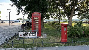 Eine rote englische Telefonzelle und ein roter englischer Briefkasten stehen neben dem Schild Greenwich Promenade auf einer Rasenfläche am Ufer.