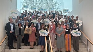 Rund siebzig Menschen stehen dicht gedrängt auf einer breiten Treppe in einem hellen Foyer und halten Schilder in Sprechblasenform mit dem Schriftzug GEM EINSAM, Allianz gegen Einsamkeit, in die Höhe.