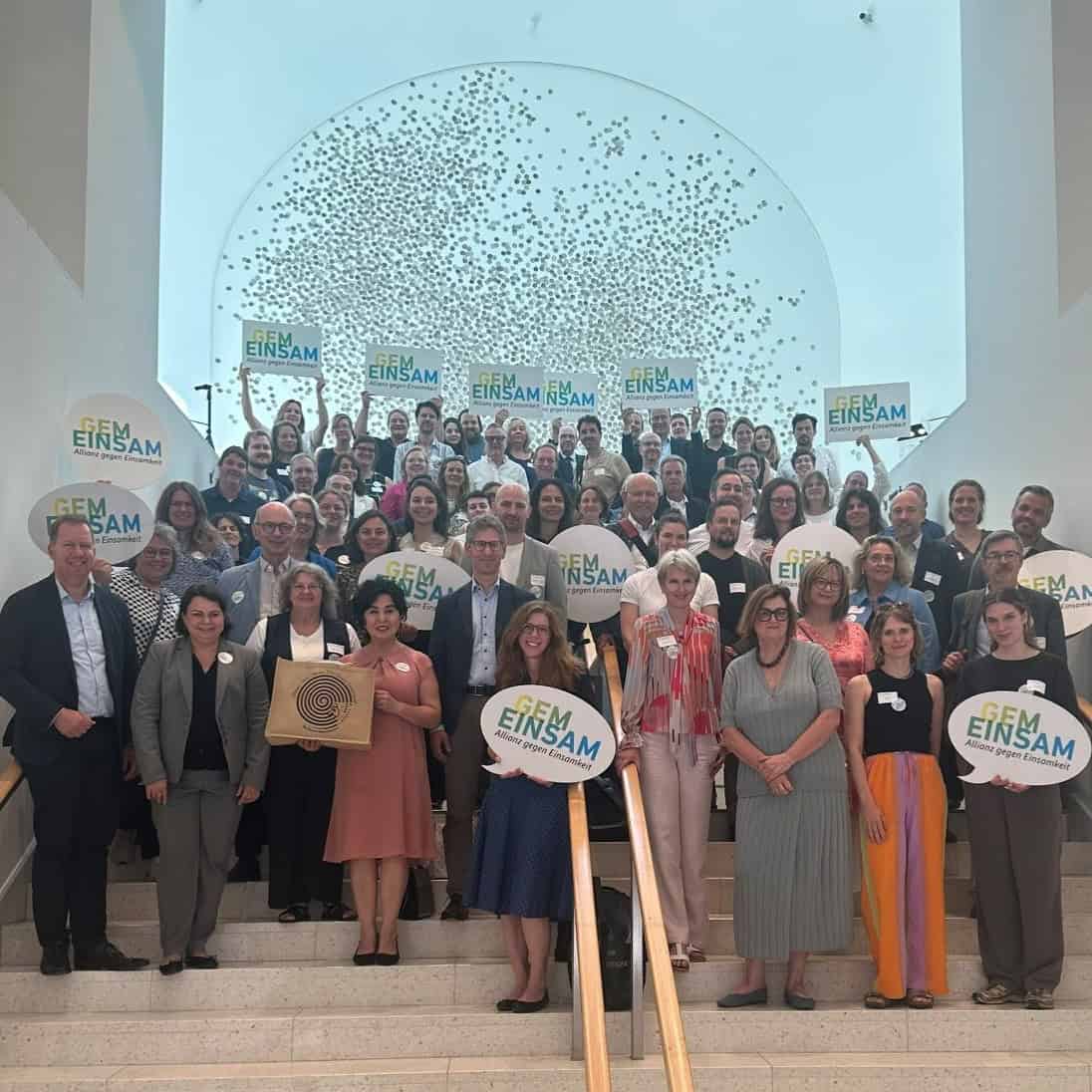Rund siebzig Menschen stehen dicht gedrängt auf einer breiten Treppe in einem hellen Foyer und halten Schilder in Sprechblasenform mit dem Schriftzug GEM EINSAM, Allianz gegen Einsamkeit, in die Höhe.