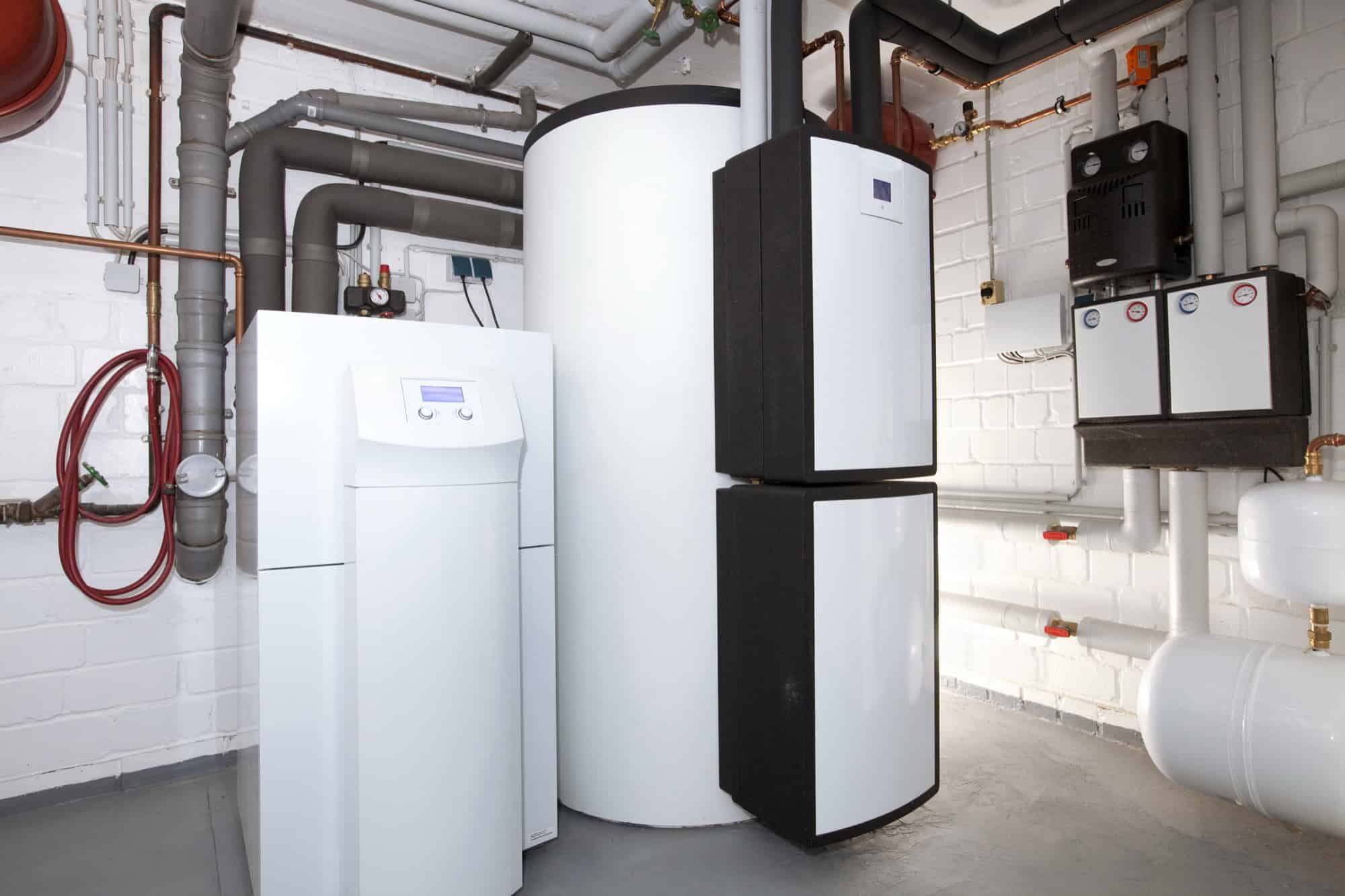 Heizungskeller mit weißen Standgeräten, einem großen Warmwasserspeicher, Kupferleitungen und Ausdehnungsgefäßen vor einer weiß gestrichenen Ziegelwand.