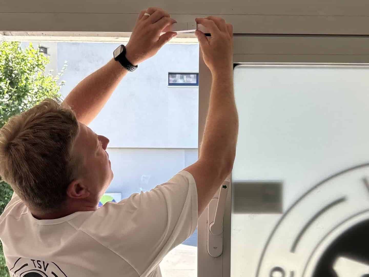 Ein Mann im weißen Shirt des TSV Berlin-Wittenau greift mit beiden Händen nach oben und setzt einen schmalen weißen Sensor an den oberen Rahmen einer Glastür.