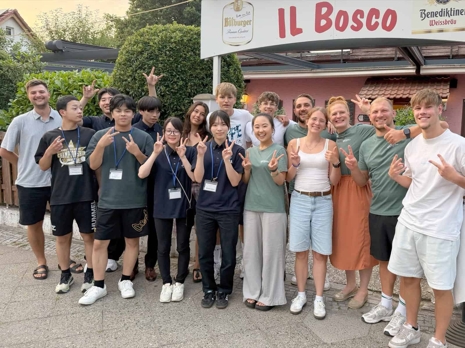 Große Gruppe Jugendlicher und Erwachsener steht am Abend vor dem Schild der Trattoria Il Bosco und zeigt das Victory-Zeichen.