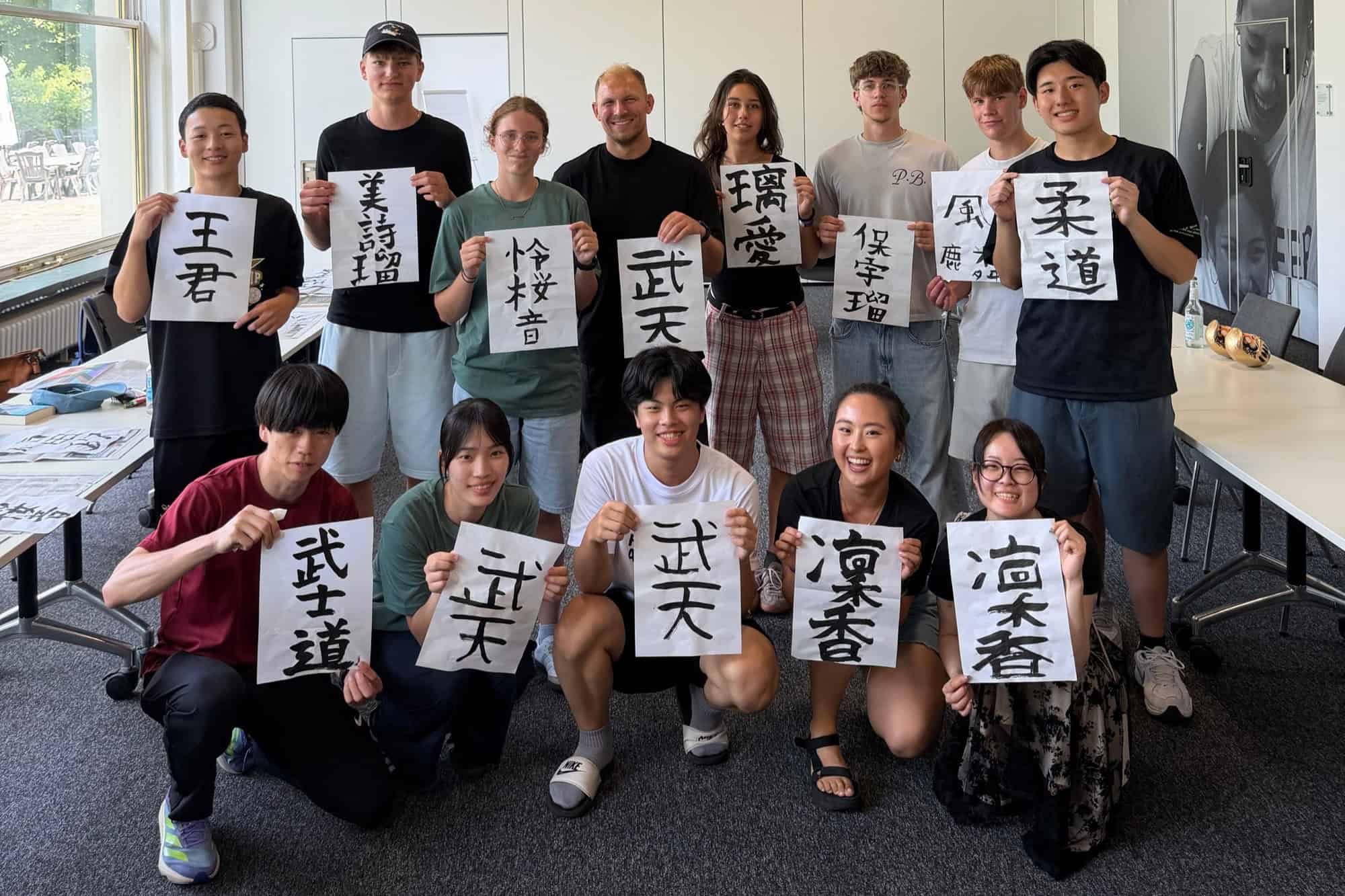 Jugendliche stehen und knien in einem hellen Seminarraum und halten Blätter mit selbst gemalten japanischen Schriftzeichen hoch.