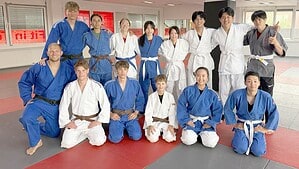 Deutsche und japanische Jugendliche in weißen und blauen Judoanzügen stehen und knien für ein Gruppenfoto auf der Judomatte.