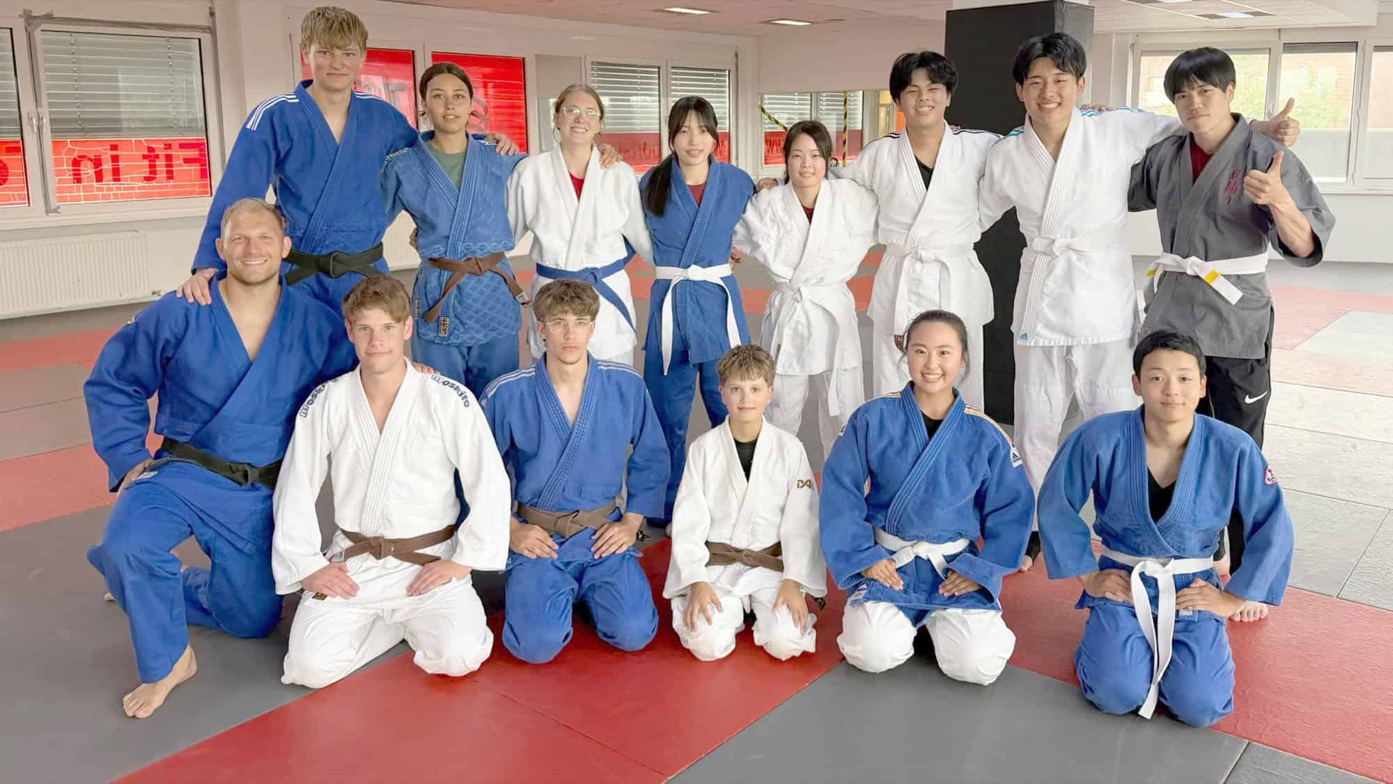 Deutsche und japanische Jugendliche in weißen und blauen Judoanzügen stehen und knien für ein Gruppenfoto auf der Judomatte.