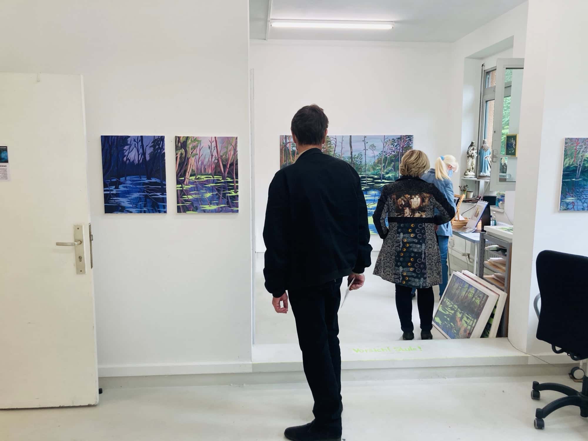 Besucherinnen und Besucher sehen sich Landschaftsbilder in einem hellen Atelier an