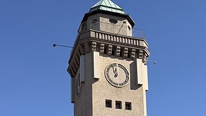 Der Kasinoturm in Frohnau vor blauem Himmel, ein hoher Turm mit heller Natursteinfassade, Turmuhr und grünem Kuppeldach, davor eine Straße mit Fußgängerüberweg und parkenden Autos.