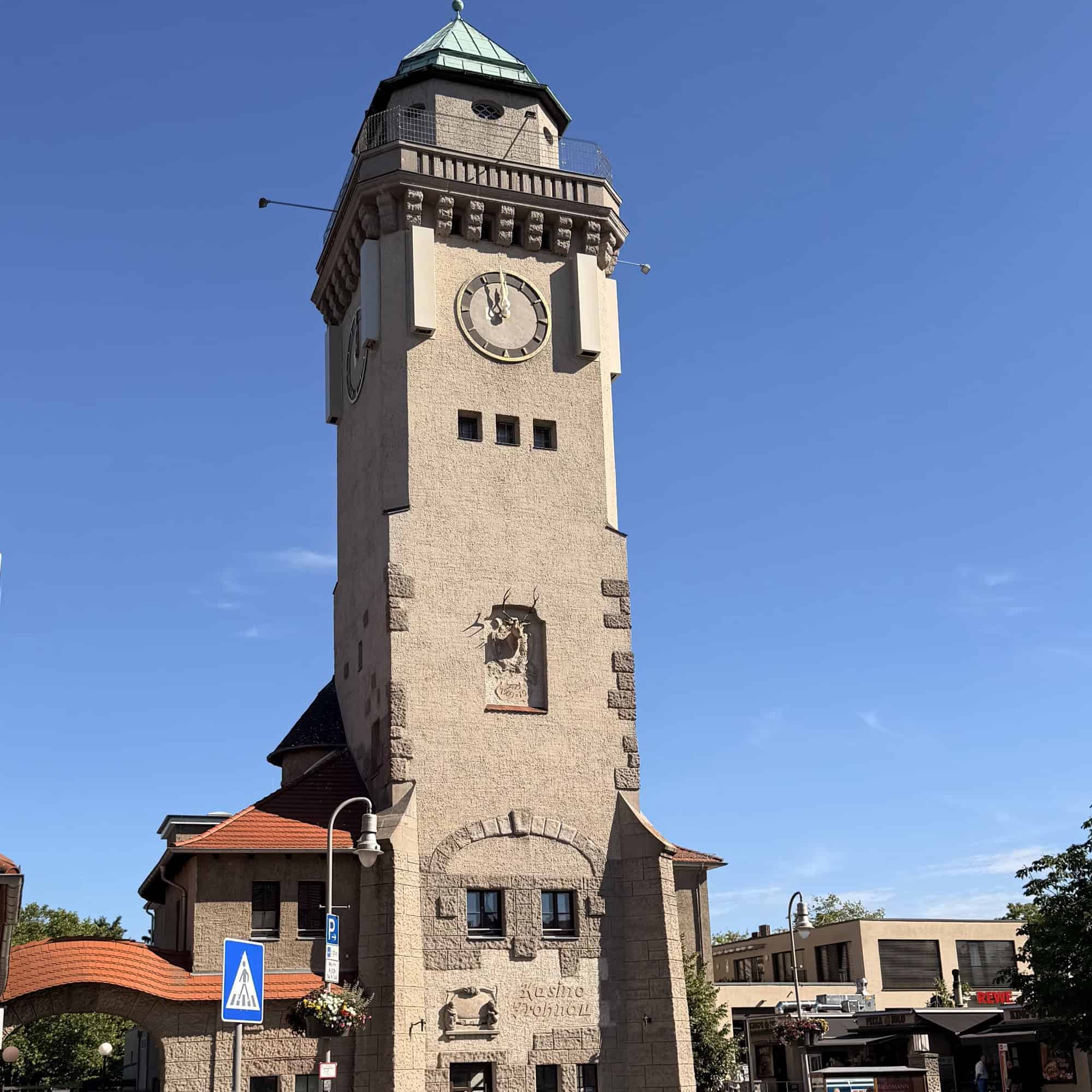 Der Kasinoturm in Frohnau vor blauem Himmel, ein hoher Turm mit heller Natursteinfassade, Turmuhr und grünem Kuppeldach, davor eine Straße mit Fußgängerüberweg und parkenden Autos.