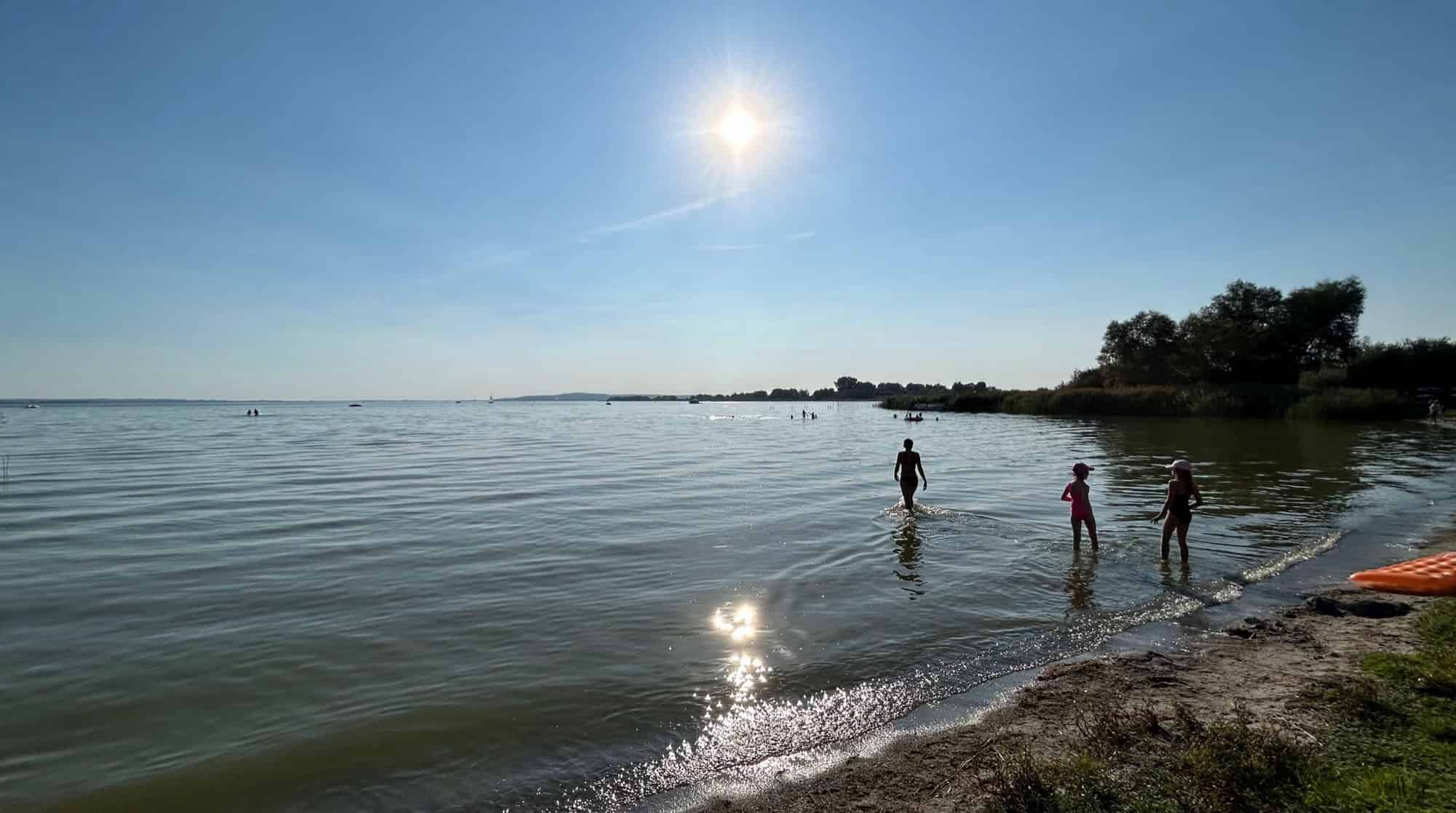 Drei Badende stehen bei hochstehender Sonne im flachen Wasser eines Sees.