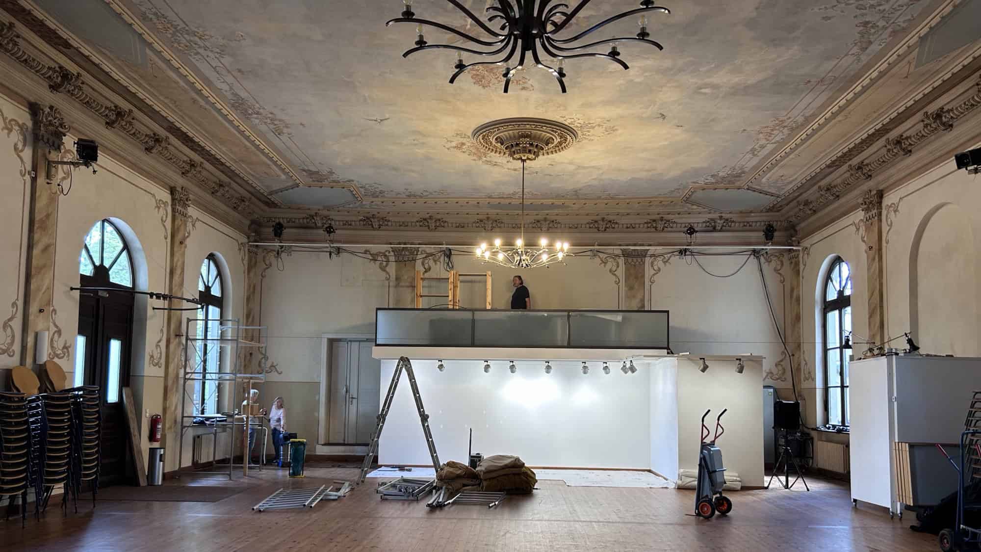 Blick in den leeren historischen Festsaal des LabSaals mit Dielenboden, zwei schmiedeeisernen Kronleuchtern, bemalter Decke und Stuckornamenten; im hinteren Teil stehen Leiter, Sackkarre und Abdeckplanen.