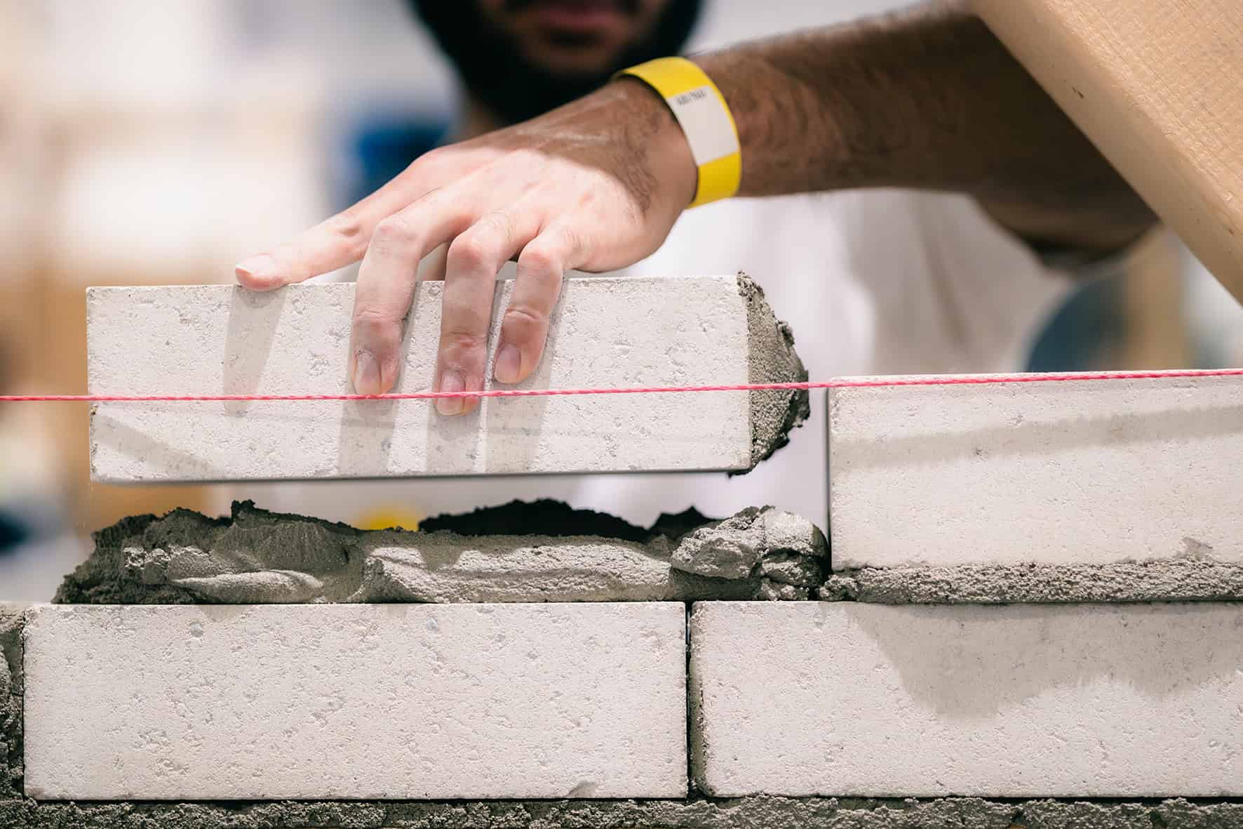 Ein junger Maurer setzt einen hellen Mauerstein an einer rot gespannten Richtschnur auf frischen Mörtel.