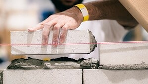 Ein junger Maurer setzt einen hellen Mauerstein an einer rot gespannten Richtschnur auf frischen Mörtel.