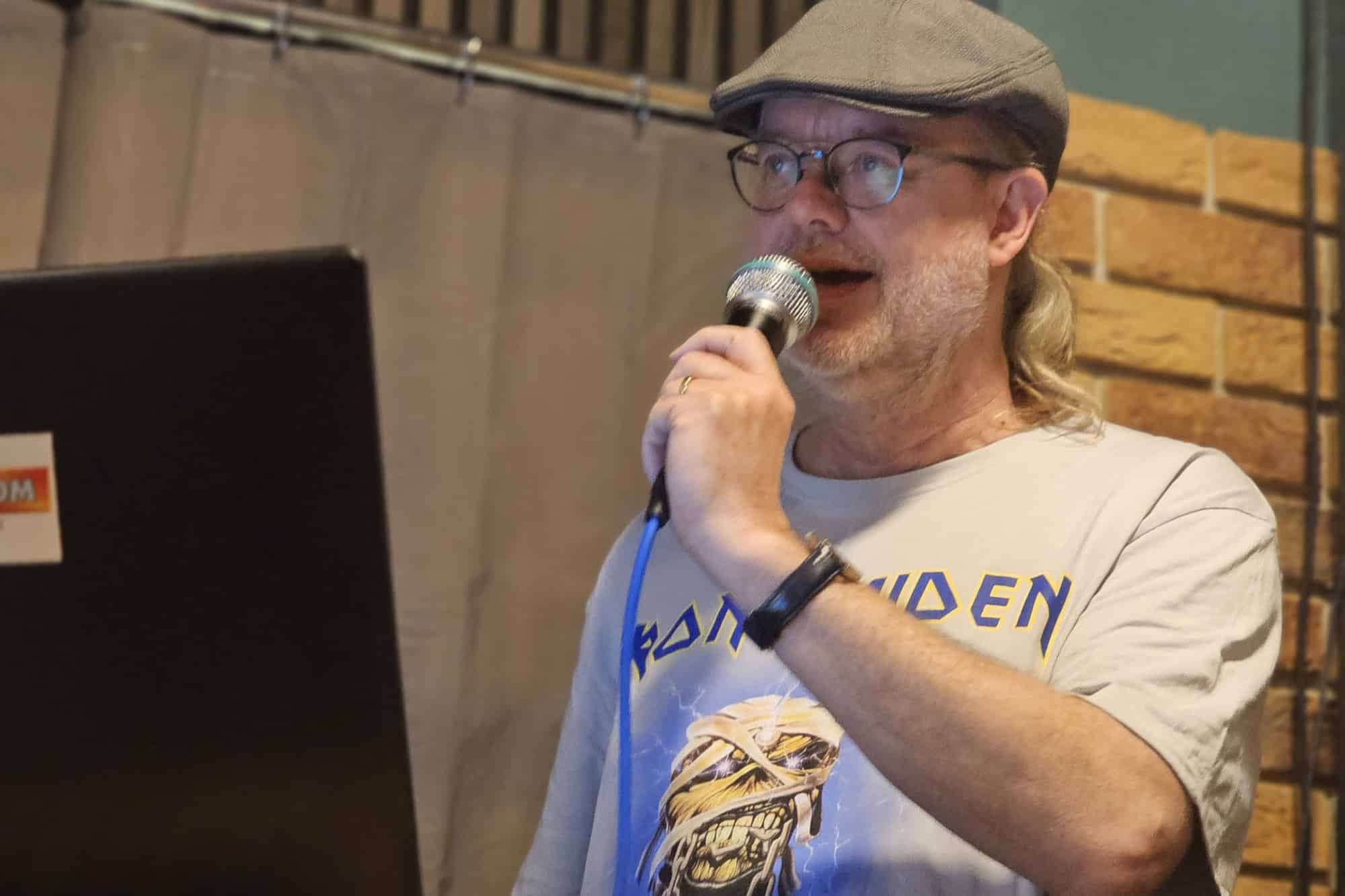 Ein Mann mit Schiebermütze, Brille und Bart spricht in ein Mikrofon, vor ihm steht ein aufgeklappter Laptop auf einem Holztresen.