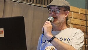 Ein Mann mit Schiebermütze, Brille und Bart spricht in ein Mikrofon, vor ihm steht ein aufgeklappter Laptop auf einem Holztresen.