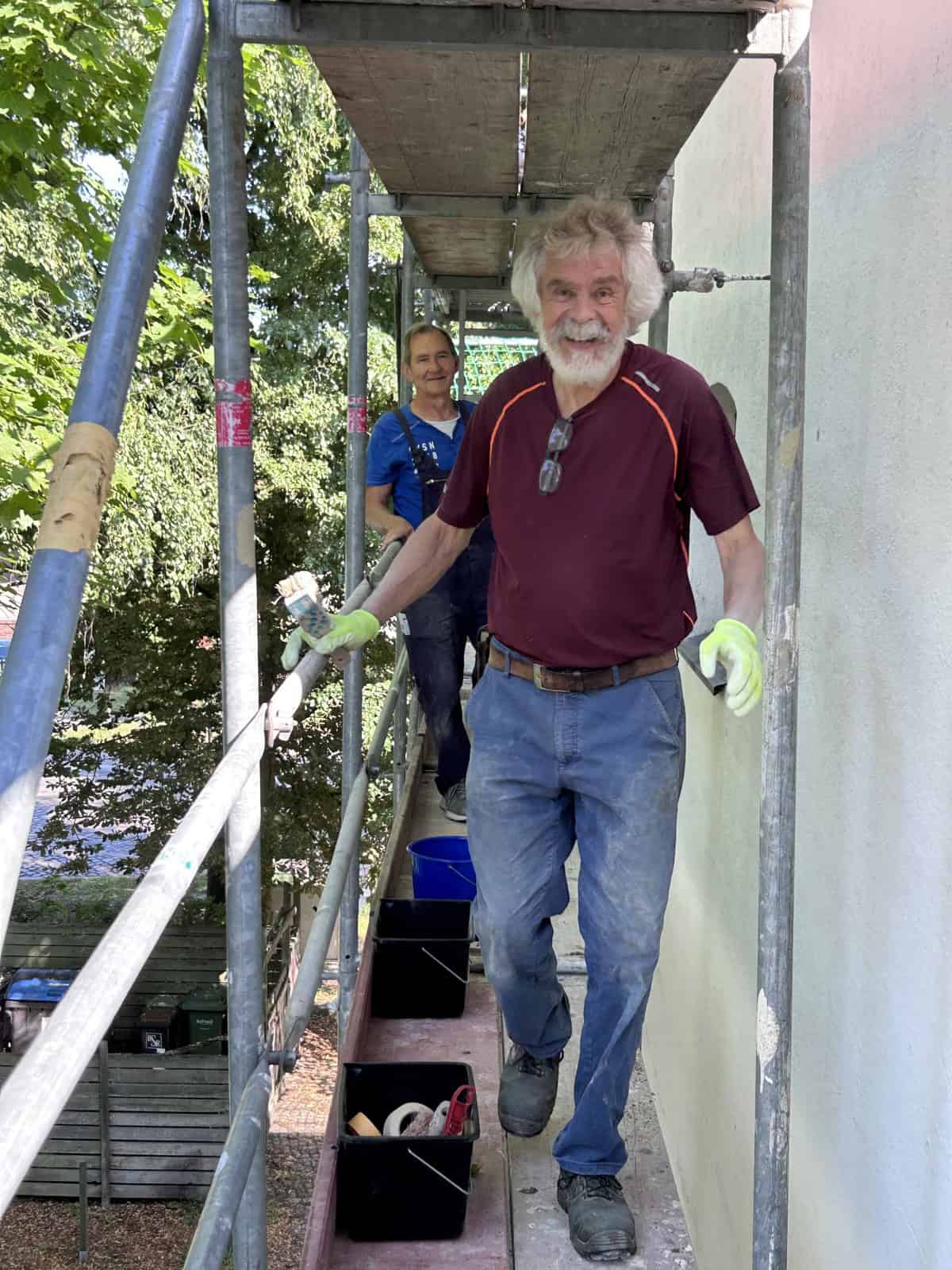 Zwei Männer stehen auf einem Baugerüst vor einer frisch verputzten Wand, der vordere mit Arbeitshandschuhen und Farbrolle, auf dem Gerüstboden stehen Eimer und Werkzeug.