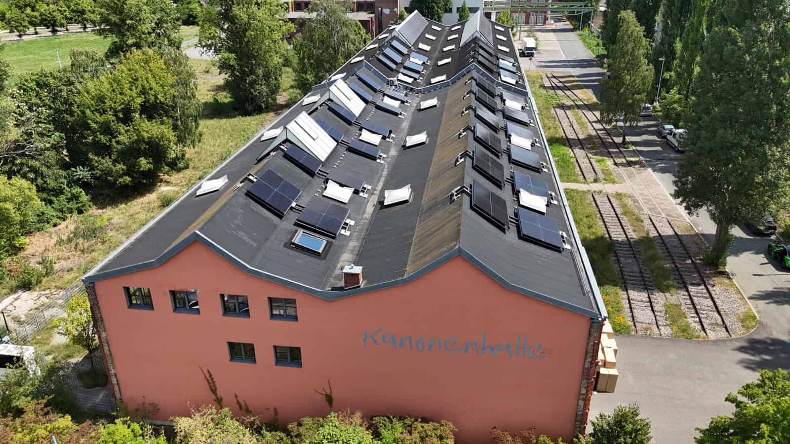 Luftaufnahme der langgestreckten Kanonenhalle mit rotem Mauerwerk, Solarmodulen auf dem Dach und Gleisen daneben.