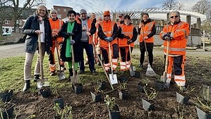 Rund ein Dutzend Menschen, viele in orangefarbener Warnkleidung, stehen mit Spaten und Rechen in einem frisch vorbereiteten Beet. Vor ihnen stehen Pflanztöpfe in Reihen, im Hintergrund die weiße Holzpergola des Zeltinger Platzes.