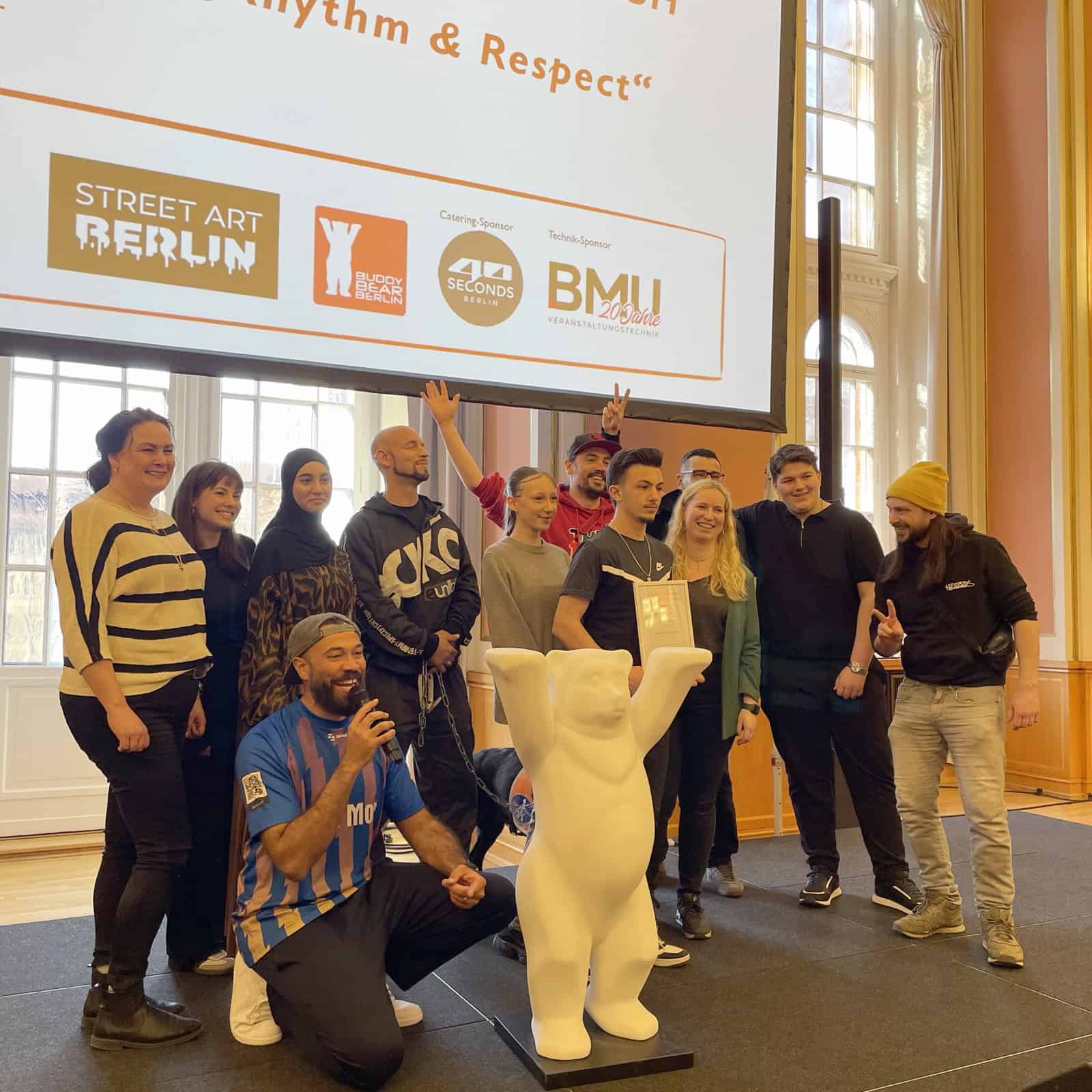 Jugendliche und Erwachsene stehen vor einer Leinwand mit der Aufschrift 1. Platz Quinoa Bildung gGmbH Rhythm & Respect