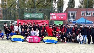 Große Gruppe von Fußballspielern, Betreuern und Gästen mit Medaillen und einem Pokal auf einem Sportplatz vor Bannern des Refugees Welcome Cup.