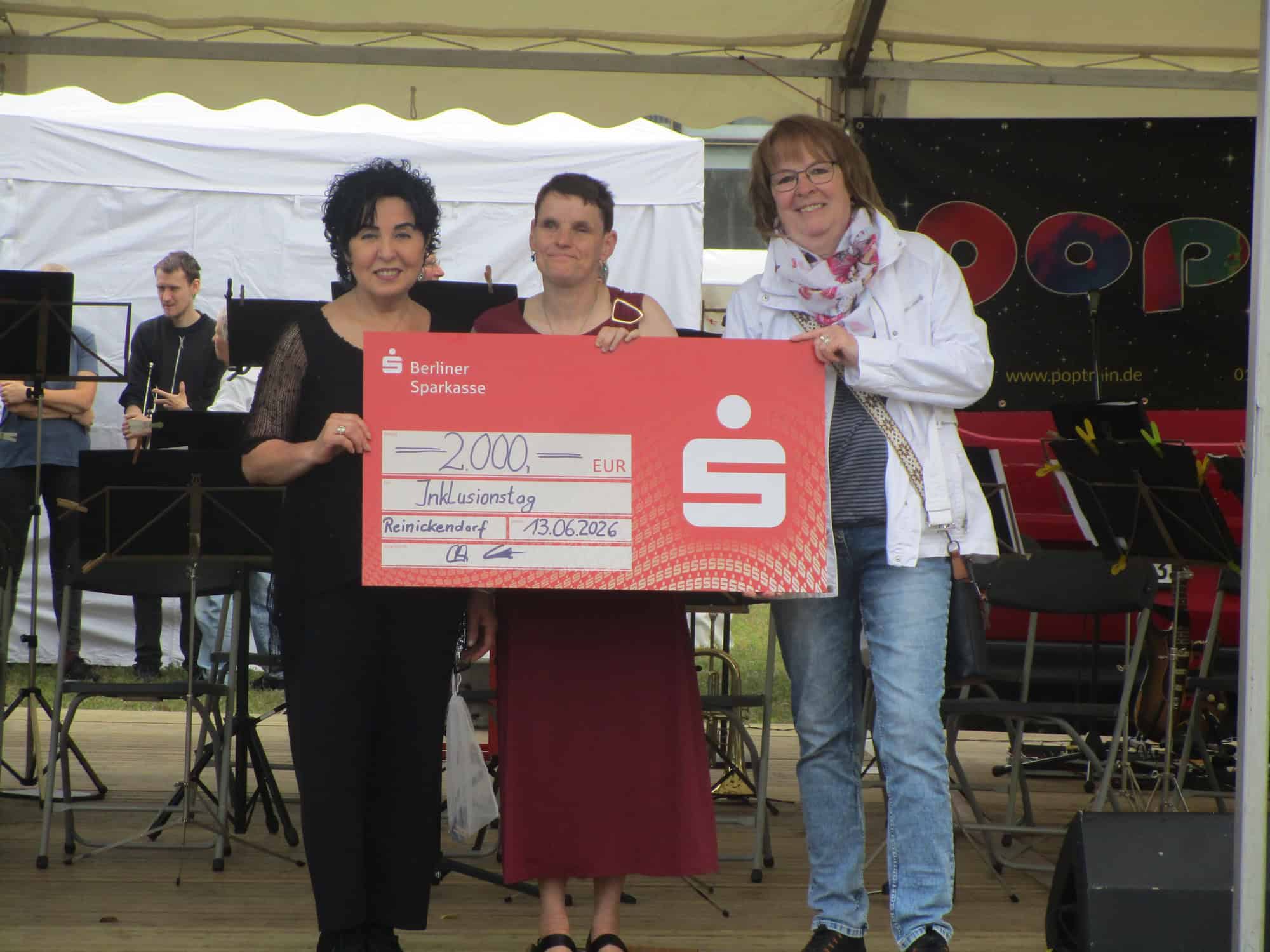 Drei Frauen stehen auf einer Bühne und halten einen großen roten Spendenscheck der Berliner Sparkasse über 2.000 Euro in die Kamera. Im Hintergrund proben Musiker eines Blasorchesters.