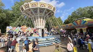 Ein buntes Kettenkarussell dreht sich vor blauem Himmel, davor stehen Besucherinnen und Besucher zwischen Buden und Fahrgeschäften.